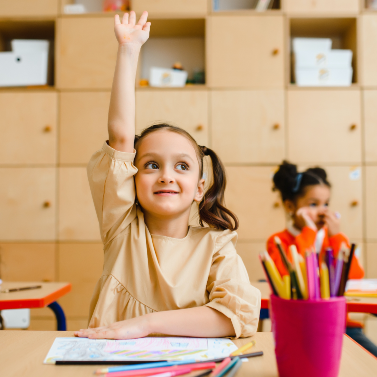 A young girl sits at a classroom desk with her hand raised.